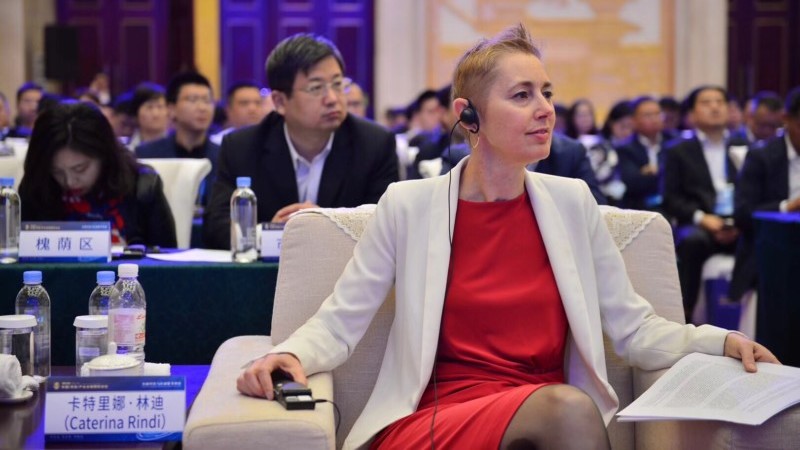Caterina Rindi in an armchair at the Jinan Forum. She is wearing a red dress with a white jacket.